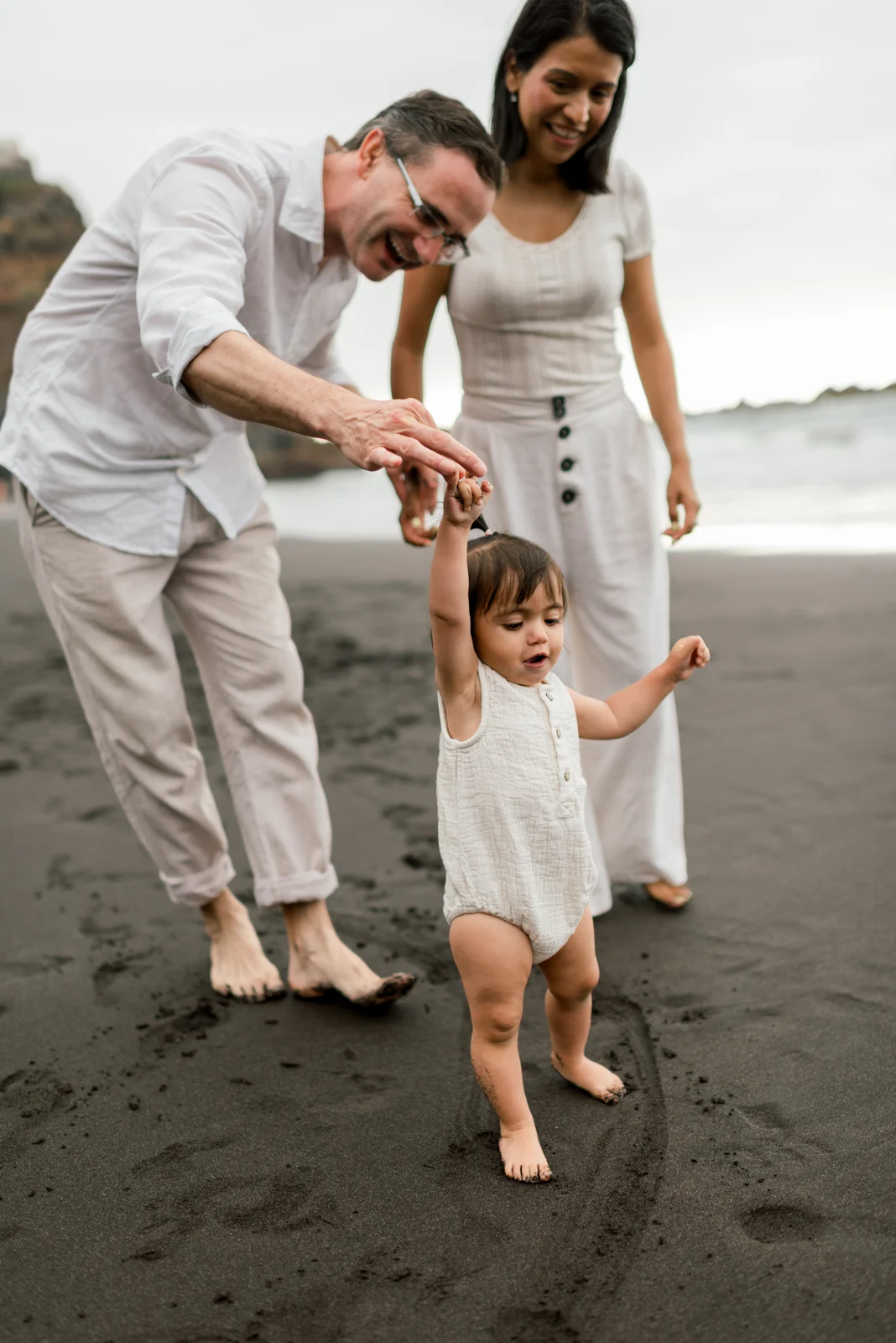A little girl walks holding her parents’ hands as they laugh beside her.