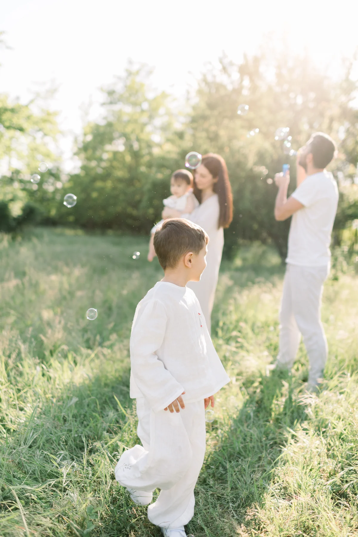 A little boy plays in tall grass while his parents and the baby watch soap bubbles.