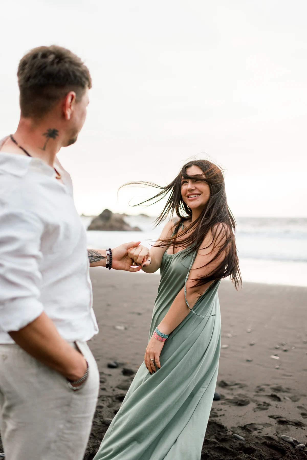 A couple holding hands and laughing on the beach, their hair and clothes moving in the wind.