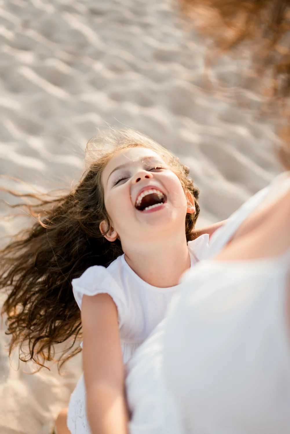 A little girl throws her head back laughing as she holds her mother’s hand on the sand.