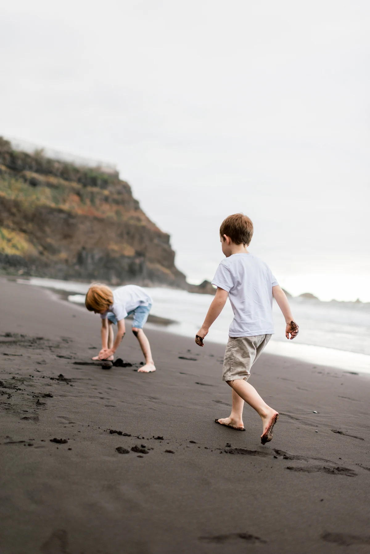 Two siblings play barefoot and explore the sand beside the water.