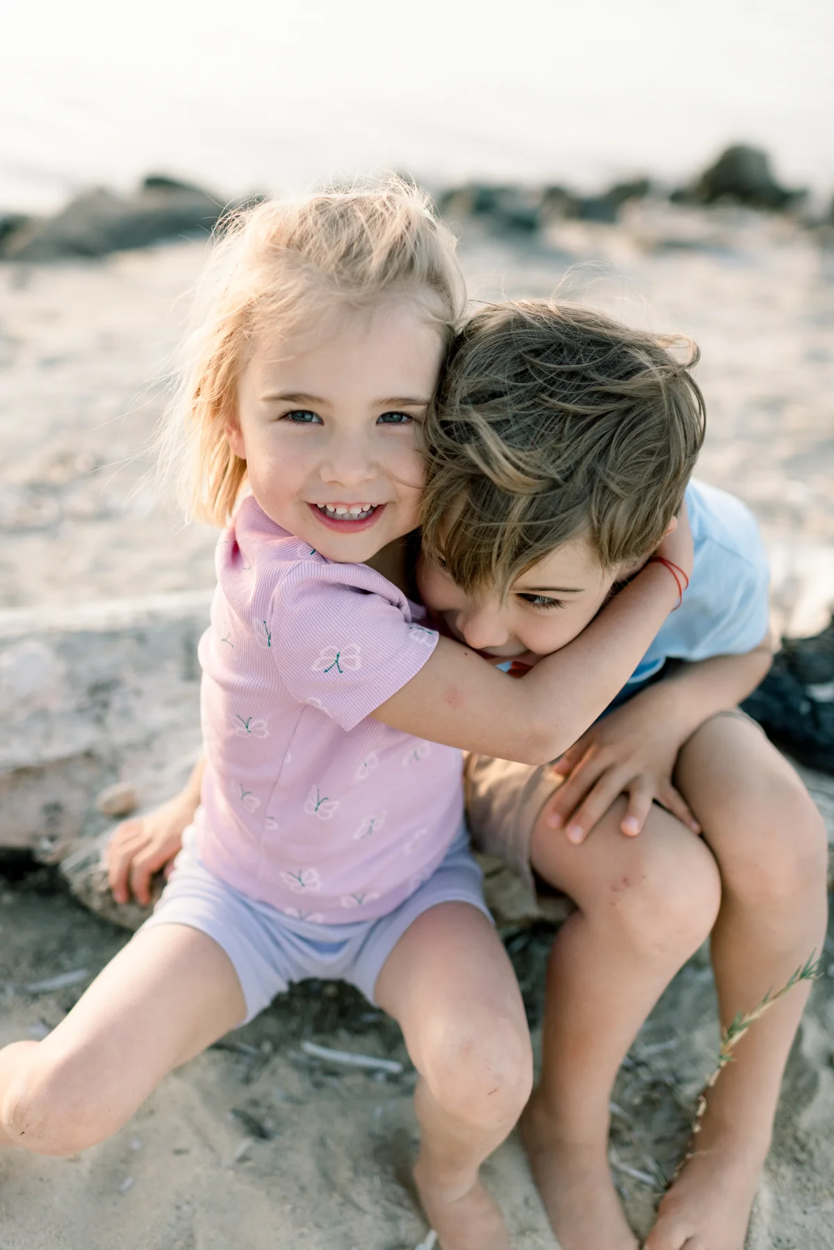 Two siblings hugging cheek to cheek on the sand.