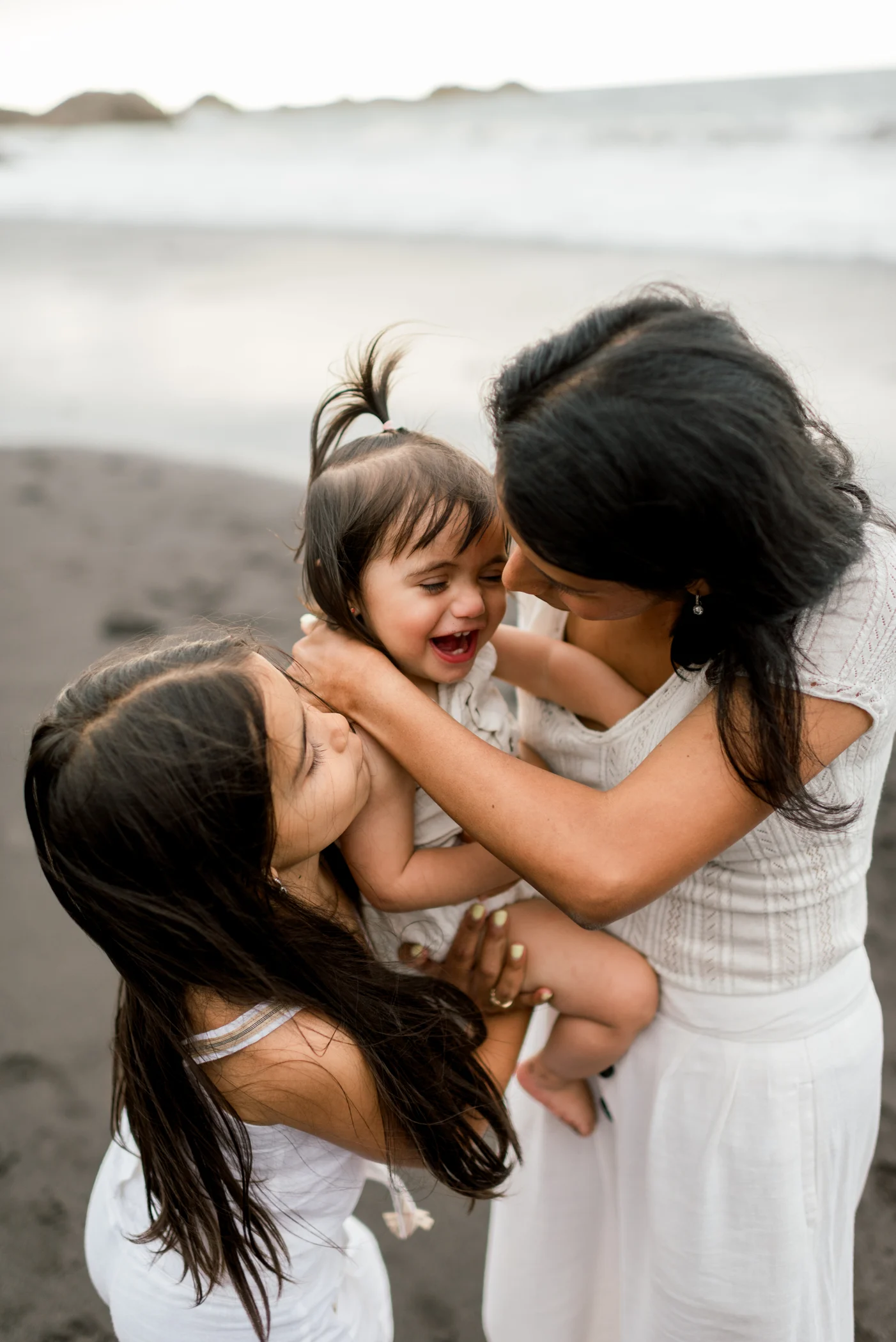 A mother hugging her two daughters as the younger one laughs in her arms.
