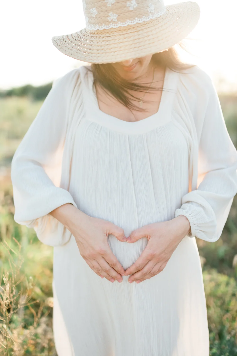 An expectant mother wearing a hat rests her hands on her bump over a white dress.