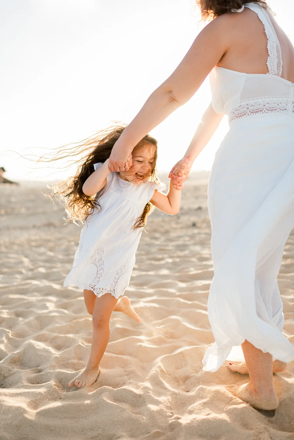 A little girl laughing as her mother holds her hands on the sand.
