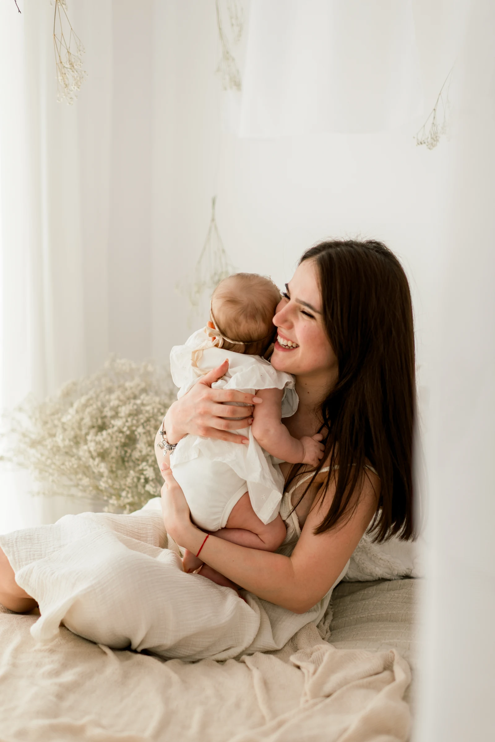 A mother smiling with her baby against her cheek on a sofa in a bright studio.