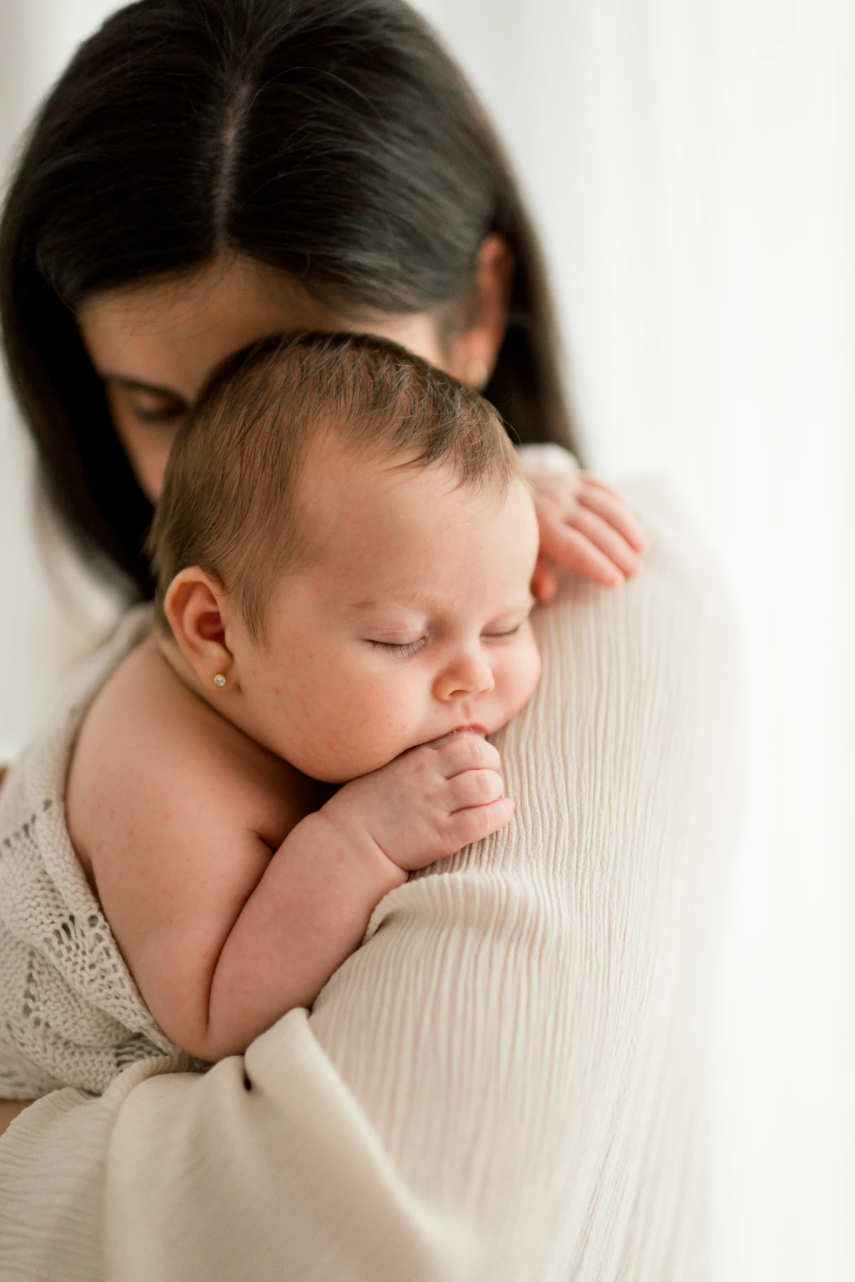 A baby sleeping on their mother’s shoulder with their hands beside their cheek.