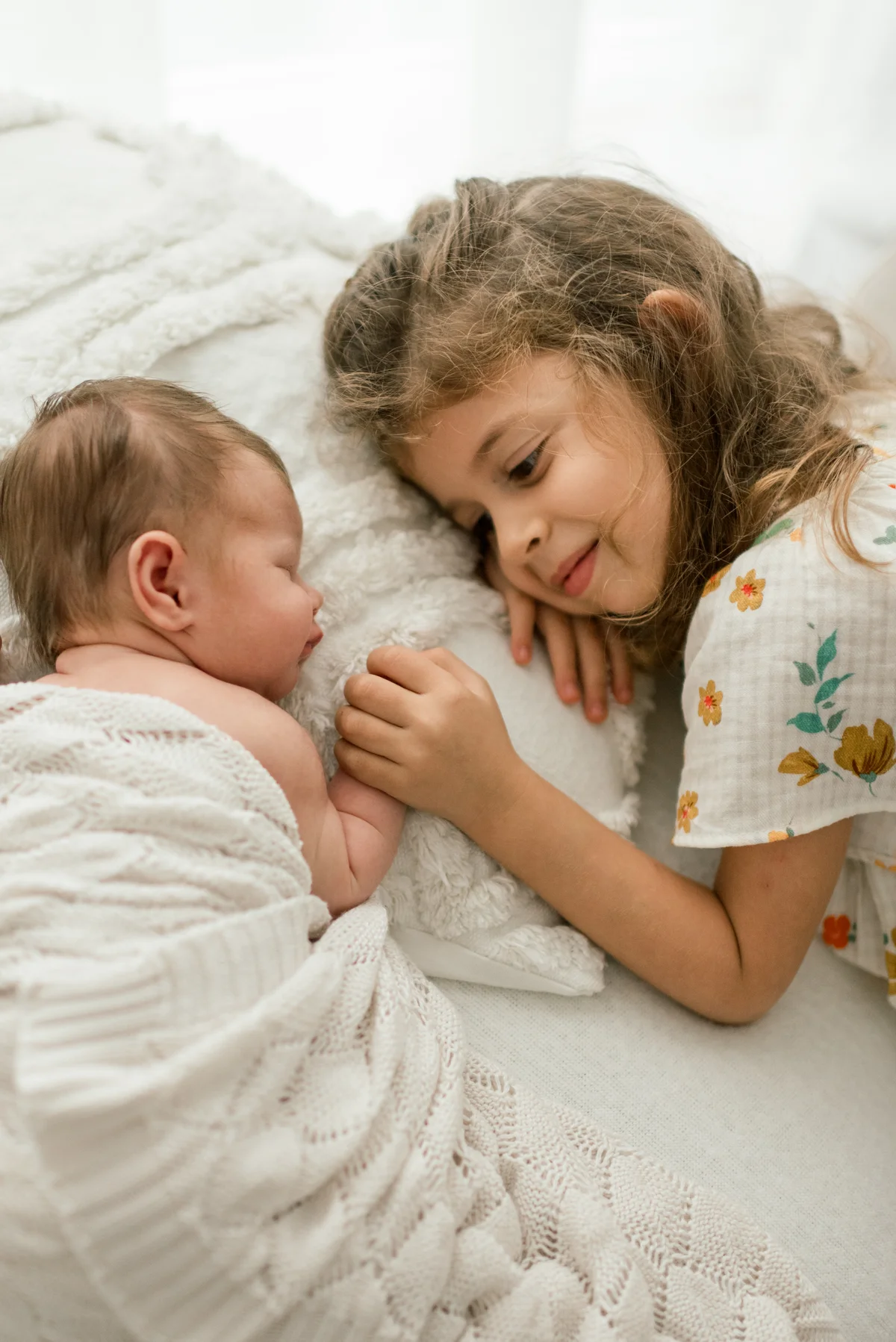 A little girl rests her cheek close to a baby wrapped in a white blanket.