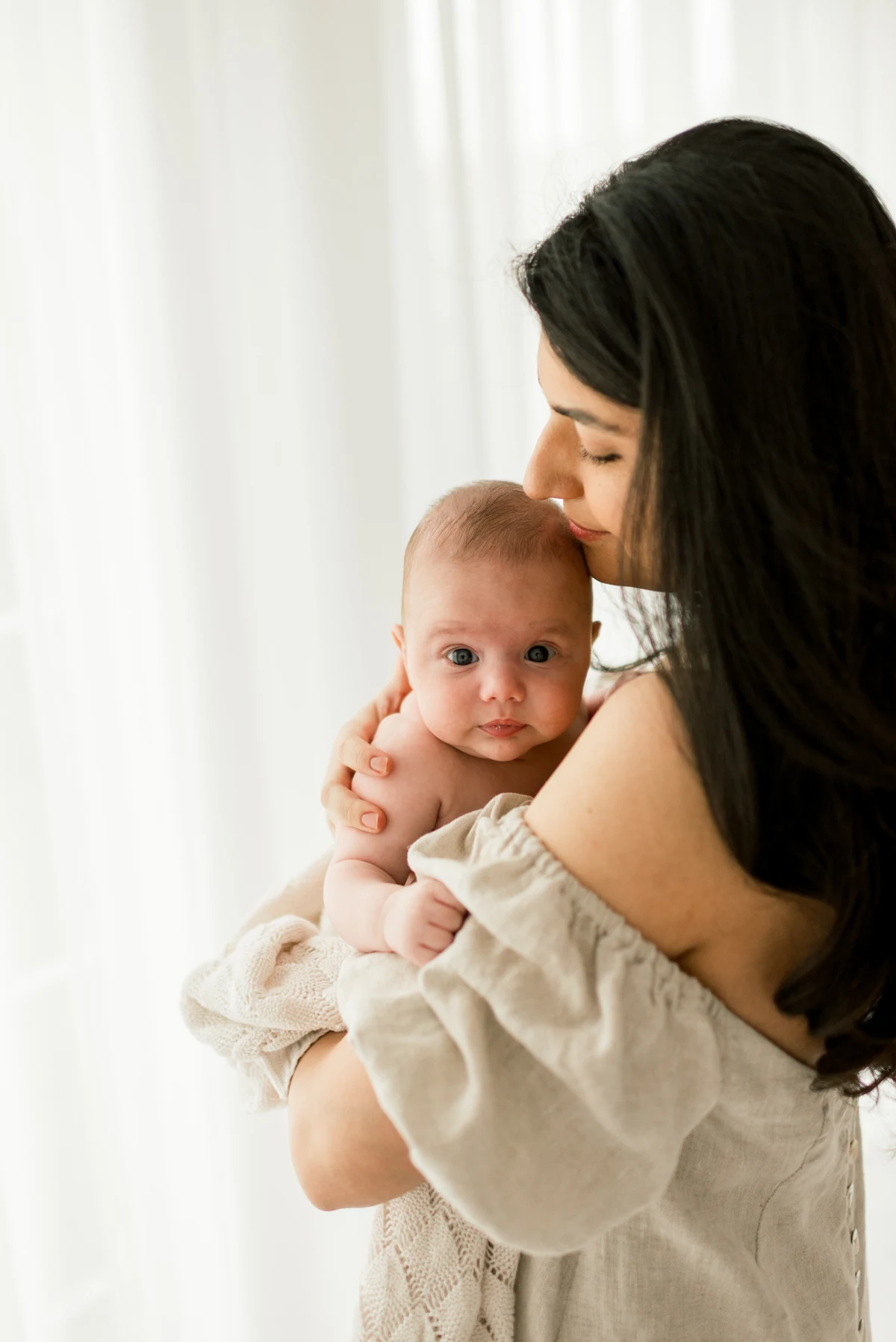 A mother kissing the top of her baby’s head, holding them beside a white curtain.
