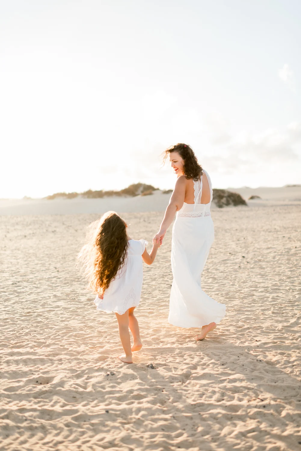 A mother and daughter walking hand in hand on the sand.